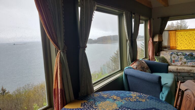 3 large windows overlooking Chiniak Bay with a round table and blue tablecloth surrounded by 2 wooden chairs.