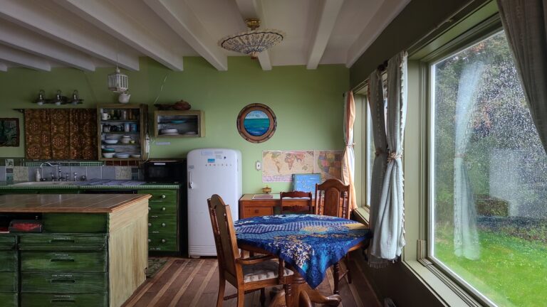 Round dining table with blue table cloth and wooden chairs overlook Chiniak Bay. There is a vintage round edged fridge against one green wall.